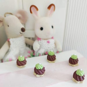 May include: A whimsical scene featuring two plush animal figures, a mouse and a rabbit, with miniature desserts. The desserts are chocolate-colored with green frosting and a silver bead on top. The scene is set on a white surface with a pink ribbon.