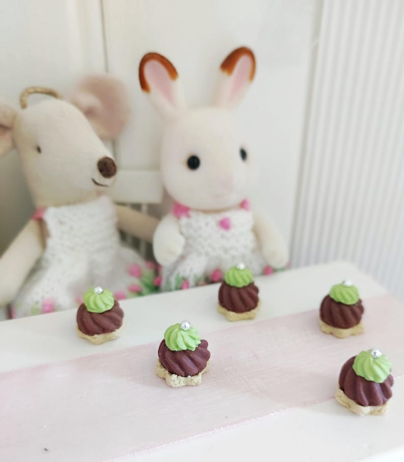 May include: A whimsical scene featuring two plush animal figures, a mouse and a rabbit, with miniature desserts. The desserts are chocolate-colored with green frosting and a silver bead on top. The scene is set on a white surface with a pink ribbon.