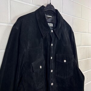 Peut inclure: Chemise en daim noir avec deux poches de poitrine et deux poches inf&eacute;rieures. La chemise a un col classique et des boutons argent&eacute;s. Elle est suspendue &agrave; un cintre noir. Style vintage.
