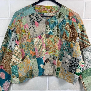 Könnte beinhalten: Eine Patchwork-Jacke mit einer Vielzahl von bunten Blumen- und geometrischen Mustern. Die Jacke hat einen runden Kragen, lange Ärmel und drei große Holzknöpfe auf der Vorderseite. Die Jacke hängt an einem schwarzen Kleiderbügel.