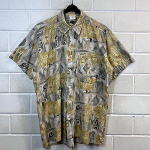 May include: A short-sleeved button-down shirt with a geometric pattern in shades of beige and gold. The shirt has a collared neckline and a button-up front.