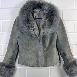Könnte beinhalten: Graue Wildlederjacke mit Kunstfellkragen und -bündchen. Die Jacke hat einen Reißverschluss und hängt an einem schwarzen Kleiderbügel. Die Jacke hat eine neutrale Farbe und scheint Vintage zu sein.