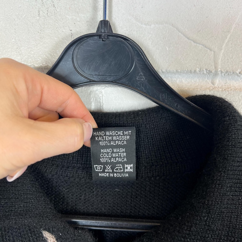 K&ouml;nnte beinhalten: Schwarzes Strickkleidungsst&uuml;ck mit einem Etikett mit der Aufschrift "HAND WASH COLD WATER 100% ALPACA MADE IN BOLIVIA". Das Etikett wird von einer Person gehalten. Das Kleidungsst&uuml;ck h&auml;ngt an einem schwarzen Kleiderb&uuml;gel.