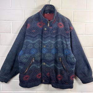 Könnte beinhalten: Dunkelblaue Fleecejacke mit einem gemusterten Design in Rot, Blau und Türkis. Die Jacke hat einen Stehkragen, einen Reißverschluss und Knopfverschlüsse. Zwei Reißverschlusstaschen sind auf der Vorderseite sichtbar.