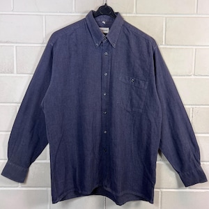 Könnte beinhalten: Ein dunkelblaues Langarmhemd mit Button-Down-Kragen und einer einzelnen Brusttasche. Das Hemd hat ein klassisches Design mit geknöpften Manschetten und hängt an einem schwarzen Kleiderbügel.