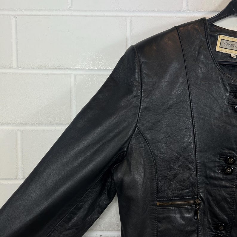 Op de afbeelding: Zwarte leren jas met een ronde kraag en knoopdetails. De jas heeft een ritszak en een label met de tekst "Soden". De jas hangt aan een zwarte hanger tegen een witte bakstenen muur.