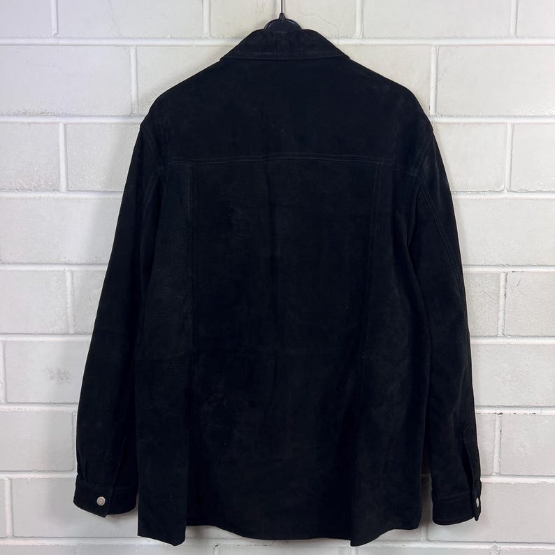 Peut inclure: Veste en daim noire avec un devant boutonn&eacute; et des manches longues. La veste est suspendue &agrave; un cintre noir contre un mur de briques blanches. La veste a un col et des poignets boutonn&eacute;s.