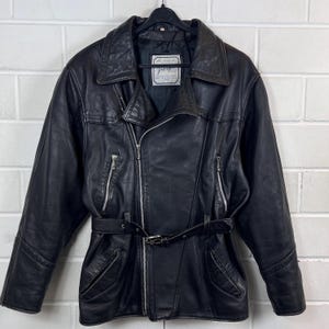 May include: Black leather jacket with a belted waist, zipper closure, and multiple pockets. The jacket has a classic biker style with a notched collar and shoulder detailing. The jacket is hanging on a black hanger against a white brick wall.
