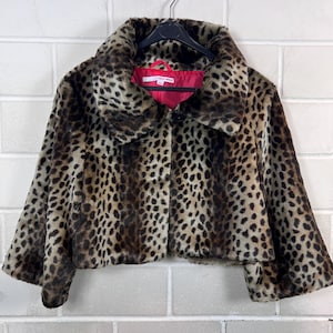 Könnte beinhalten: Kurze Jacke aus Kunstfell mit Leopardenmuster. Die Jacke hat einen breiten Kragen und ein rotes Futter. Sie hängt an einem schwarzen Kleiderbügel vor einer weißen Backsteinmauer.