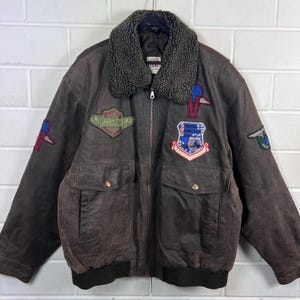 Könnte beinhalten: Eine dunkelbraune Leder-Bomberjacke mit einem dunkelgrauen Kunstfellkragen, einem Frontreißverschluss und zwei großen Pattentaschen. Die Jacke ist mit verschiedenen gestickten Aufnähern auf der Brust und den Ärmeln verziert, darunter ein schildförmiger Aufnäher.