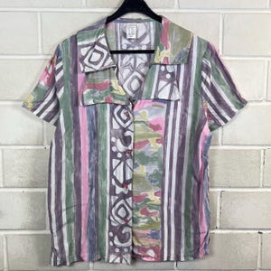 Könnte beinhalten: Kurzärmeliges Hemd mit farbenfrohem Streifenmuster. Das Hemd hat vertikale Streifen in Lila, Rosa, Grün und Weiß, mit dekorativen geometrischen und abstrakten Designs. Kragen und Knöpfe sind sichtbar.