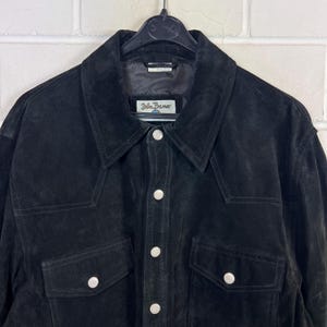 Peut inclure: Veste en daim noire avec une fermeture boutonn&eacute;e, deux poches de poitrine et un col classique. La veste porte une &eacute;tiquette "John Banner". La veste est suspendue &agrave; un cintre noir.