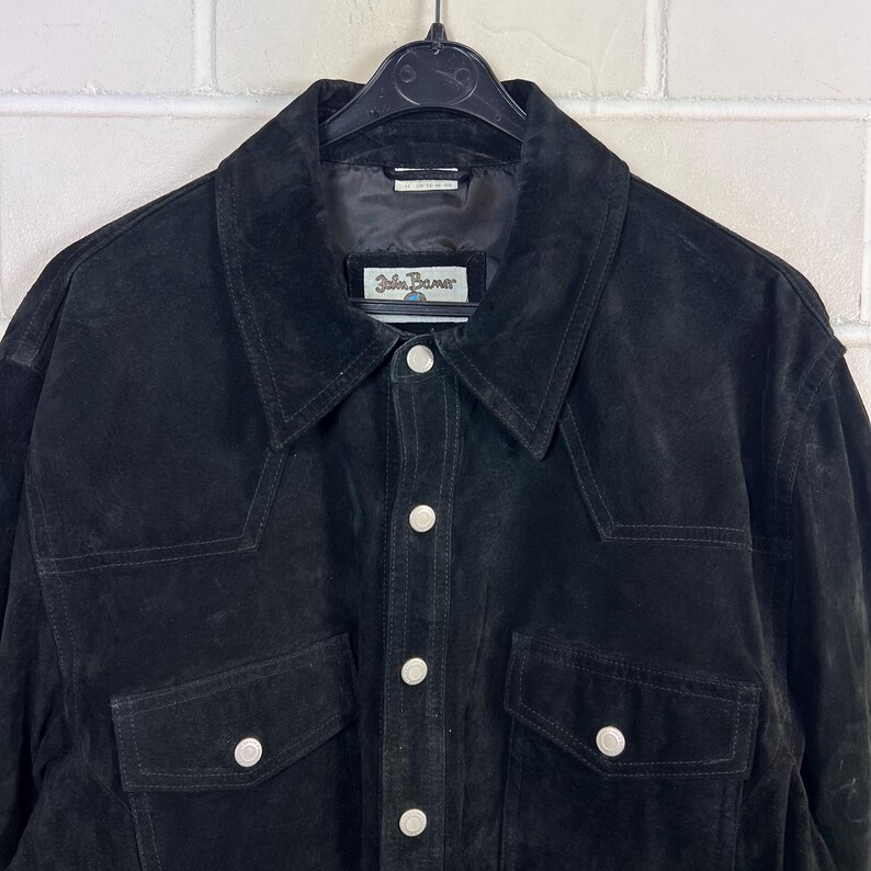 Peut inclure: Veste en daim noire avec une fermeture boutonn&eacute;e, deux poches de poitrine et un col classique. La veste porte une &eacute;tiquette "John Banner". La veste est suspendue &agrave; un cintre noir.