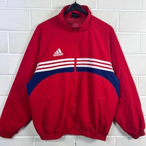 May include: Red Adidas track jacket with white stripes and blue detailing. It has a full zip, a stand-up collar, and the Adidas logo on the chest. The jacket includes two front pockets and ribbed cuffs.