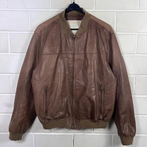May include: A brown leather bomber jacket featuring a full-length front zipper, a ribbed collar, ribbed cuffs, and a ribbed hem. The jacket includes two vertical zippered pockets on the front and horizontal seam detailing across the chest area.