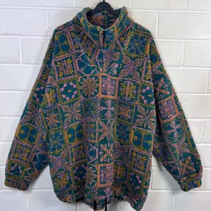 Könnte beinhalten: Ein langärmeliger Fleece-Pullover mit hohem Kragen und kurzem Reißverschluss. Das Kleidungsstück zeigt ein sich wiederholendes quadratisches Gittermuster in den Farben Petrol, Pink und Senfgelb, mit Kordelzug am Saum und gerippten Ärmelbündchen.