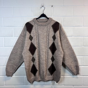 May include: A beige argyle sweater featuring brown and olive green diamond shapes. The sweater has a crew neck and cable knit detailing. It hangs on a black hanger against a white brick wall.