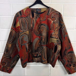 Könnte beinhalten: Eine langärmelige Jacke mit einem auffälligen, abstrakten Druck in den Farben gebranntes Orange, Dunkelbraun und mattes Gold. Das Kleidungsstück hat einen Rundhalsausschnitt, eine Knopfleiste vorne mit vier sichtbaren Knöpfen und einen geraden Saum.