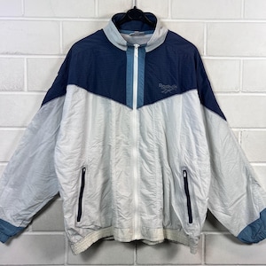 May include: A vintage Reebok windbreaker jacket in white, navy blue, and light blue. The jacket features a full zip, two front pockets, and the Reebok logo. It hangs on a black hanger against a white brick wall.