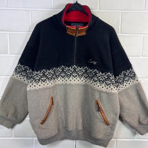 May include: A black, grey, and beige fleece pullover with a red collar lining. The top section is black, with a beige and black geometric pattern. The lower section is grey, with two zippered pockets trimmed in brown leather. The word "Sluk" is embroidered on the front.