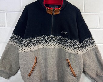 May include: A black, gray, and beige fleece pullover with a red collar lining. The top portion is black, with a beige and black geometric pattern. The lower portion is gray, with two zippered pockets trimmed in brown leather. The word "Sluk" is embroidered on the front.