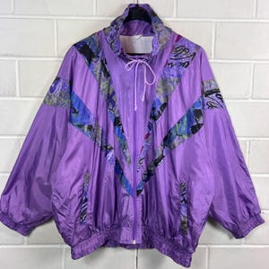 Puede incluir: Una chaqueta cortavientos de color morado con cuello alto y cordón ajustable, cremallera frontal completa y mangas largas con puños elásticos. La prenda presenta un patrón en forma de V con detalles gráficos abstractos en azul y negro.