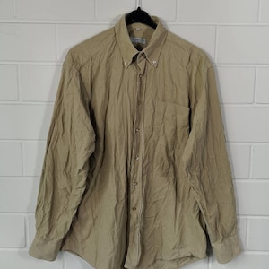 Può includere: Una camicia beige a maniche lunghe con bottoni e una tasca sul petto. La camicia è realizzata in tessuto strutturato e ha un aspetto leggermente stropicciato.