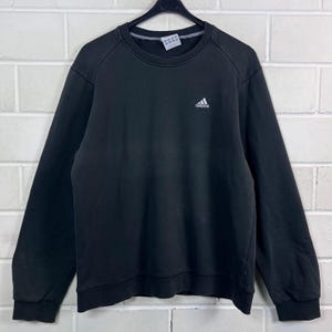 Könnte beinhalten: Ein schwarzes Langarm-Sweatshirt mit Rundhalsausschnitt und einem kleinen, weißen, aufgestickten Adidas-Logo auf der linken Brust. Das Kleidungsstück hat gerippte Bündchen und einen gerippten Saum sowie ein sichtbares Innenetikett und einen schwarzen Kleiderbügel.
