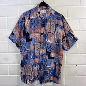 Könnte beinhalten: Kurzärmeliges Hemd mit farbenfrohem, abstraktem Muster. Das Hemd zeigt eine Mischung aus blauen, roten, schwarzen und weißen Formen mit einem sich wiederholenden Design. Das Hemd hängt an einem schwarzen Kleiderbügel.