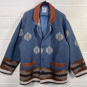 Op de afbeelding: Een blauwe jas met een bruine kraag en zakafwerking, voorzien van een geometrisch patroon in beige en bruin. De jas heeft drie blauwe knopen aan de voorzijde en een label in de kraag met de tekst GDC's. De zoom en mouwen hebben een brede band.