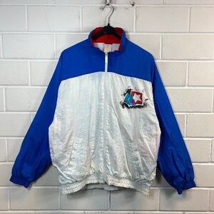 May include: A white and blue windbreaker jacket with a red zip and a logo on the left chest. The logo is a star with a red, white and blue design.