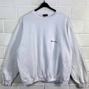 May include: A white Champion sweatshirt with a crew neck and long sleeves. The Champion logo is embroidered in red and navy blue on the chest. The sweatshirt is hanging on a black hanger.
