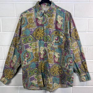 Könnte beinhalten: Ein langärmeliges Hemd mit farbenfrohem Muster. Das Hemd zeigt eine Collage aus geometrischen Formen, floralen Motiven und Wirbeln in Türkis, Lila, Gold und Grün. Es hängt an einem schwarzen Kleiderbügel vor einer weißen Backsteinmauer.