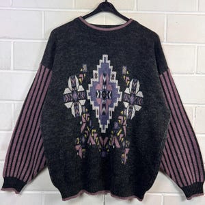 Könnte beinhalten: Ein anthrazitfarbener Langarmpullover mit einem markanten zentralen geometrischen Muster in Lavendel, Weiß und Gelb. Die Ärmel zeigen vertikale Streifen in Lavendel und Anthrazit, ergänzt durch einen lavendelfarbenen Saum am Rundhalsausschnitt.