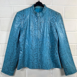 Könnte beinhalten: Türkisblaue Jacke mit Schlangenmuster. Die Jacke hat einen Stehkragen und einen durchgehenden Reißverschluss. Die Jacke hängt an einem schwarzen Kleiderbügel vor einer weißen Backsteinmauer.