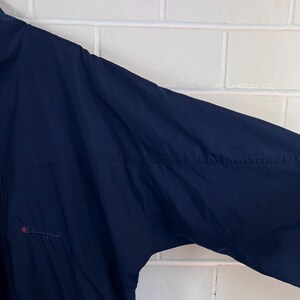 May include: Navy blue Champion jacket with a full zipper and the Champion logo embroidered in red on the chest. The jacket is made of a lightweight material, suitable for layering. The jacket's design is simple and classic.