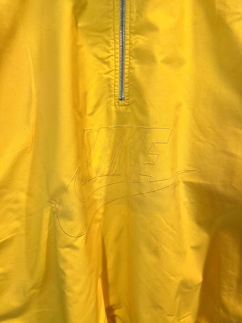 Puede incluir: Una chaqueta cortavientos amarilla con cremallera y un gran logotipo de Nike bordado en la parte delantera.