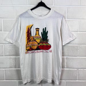 Könnte beinhalten: Weißes T-Shirt mit einem farbenfrohen Grafikdesign, das Töpferwaren, einen Kaktus und Chilischoten zeigt. Der Text "San Antonio TX." ist unter dem Bild aufgedruckt. Das Design verwendet Gelb-, Rot-, Grün- und Brauntöne.
