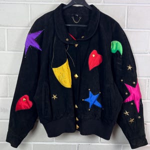 Può includere: Giacca in camoscio nera con applicazioni colorate a forma di stelle e cuori. Presenta un colletto rotondo, borchie dorate e orlo a coste. Design artistico unico con accenti viola, rossi, blu, verdi e gialli.