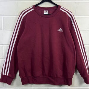 Könnte beinhalten: Burgunderfarbenes Adidas-Sweatshirt mit weißen Streifen auf den Ärmeln. Das Adidas-Logo befindet sich auf der Brust. Das Sweatshirt hat einen Rundhalsausschnitt und gerippte Bündchen und Saum.