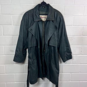 Könnte beinhalten: Ein dunkelgrüner Ledertrenchcoat mit breitem Kragen, Frontreißverschluss und Gürtel. Das Innenetikett zeigt den Text Torino Collection. Der Mantel hat lange Ärmel, eine Sturmklappe und zwei Vordertaschen.
