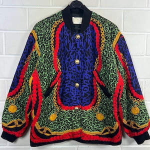Könnte beinhalten: Vintage Bomberjacke mit auffälligem Design. Die Jacke hat einen schwarzen Kragen und Manschetten, goldene Knöpfe und einen Mix aus Mustern. Leopardendruck in Grün und Schwarz, Blau, Rot und goldene Kettenakzente erzeugen einen auffälligen Look.