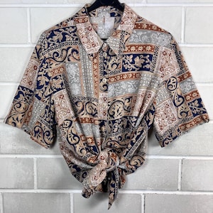May include: Short-sleeved shirt with a button-down front and a knotted tie. The shirt displays a repeating pattern of navy blue, tan, brown, and grey geometric and floral designs. It hangs on a black hanger against a white brick wall.
