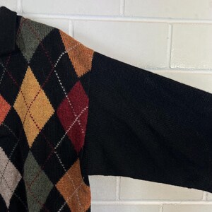 K&ouml;nnte beinhalten: Ein schwarzer Pullover mit einem Rautenmuster in Orange-, Gelb-, Rot- und Gr&uuml;nt&ouml;nen. Die &Auml;rmel sind einfarbig schwarz. Der Pullover hat einen Kragen.