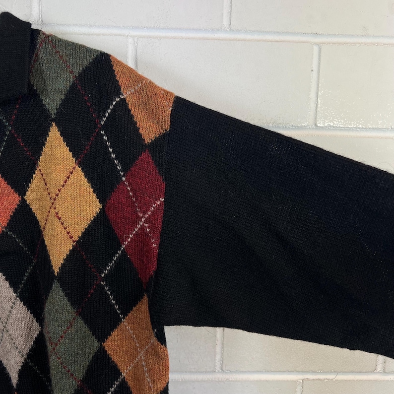 K&ouml;nnte beinhalten: Ein schwarzer Pullover mit einem Rautenmuster in Orange-, Gelb-, Rot- und Gr&uuml;nt&ouml;nen. Die &Auml;rmel sind einfarbig schwarz. Der Pullover hat einen Kragen.