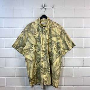 May include: A yellow short-sleeved button-down shirt with a green and brown abstract leaf pattern. The shirt has a collar and a button closure.