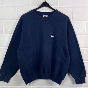 Pode incluir: Uma camisola de manga comprida e gola redonda em azul-marinho com um pequeno logótipo Nike swoosh bordado a branco no lado esquerdo do peito. A peça tem punhos e bainha canelados, com uma etiqueta da marca Nike visível no interior.