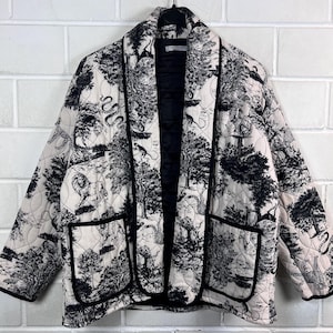 May include: A quilted jacket featuring a black and white forest scene print. The jacket has a black lining, black trim, and two front pockets. The design includes trees, animals, and a diamond pattern.