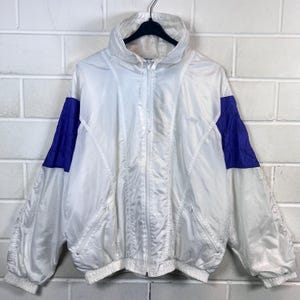 Pode incluir: Casaco branco vintage com painéis azuis nos ombros, fecho de correr completo e gola alta. O casaco está pendurado num cabide preto. Fundo: parede de tijolos brancos.