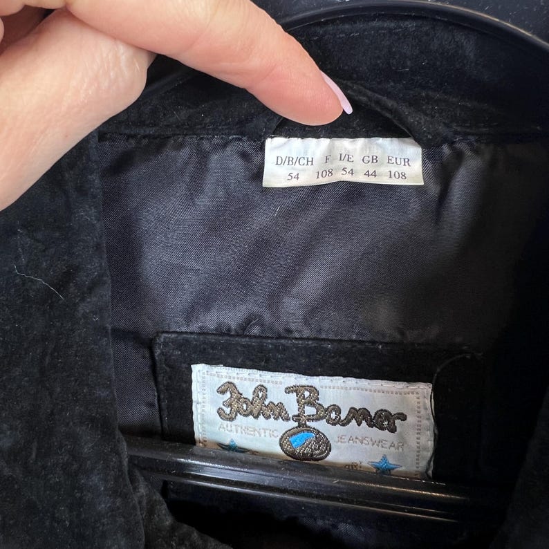 Peut inclure: Veste en daim noire avec une doublure en satin. L'&eacute;tiquette indique "John Baner Authentic Jeanswear" et comprend des informations de taille. La veste est suspendue &agrave; un cintre noir.